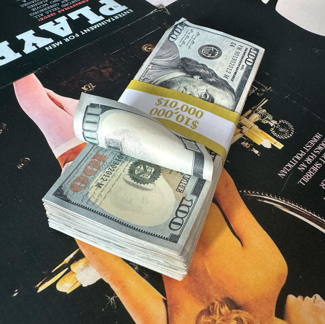 A stack of prop money designed to look like US $10,000 bills, featuring a weathered appearance and bundled with a bank strap, placed on a surface with movie posters in the background.
