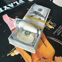 A stack of prop money designed to look like US $10,000 bills, featuring a weathered appearance and bundled with a bank strap, placed on a surface with movie posters in the background.
